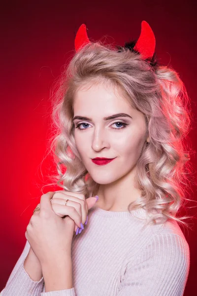 Portrait of a beautiful blonde woman with a cunning look and horns on her head. The dress for the Halloween party. To think something is not good. Evil demon girl is the devil. Red background