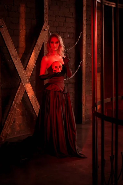 Beautiful, sexy young woman in a black evening dress in red light on a background of a cross and a skull of a man in hands. Sexy girl from hell.