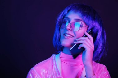 Neon ışıklı şık bir genç kadın elinde bir akıllı telefon tutuyor. Kız peruk, gözlük ve parlak bir halo ceket giyiyor. Boşluğu kopyala.