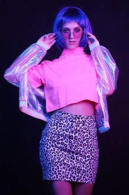 Bir gece kulübünde neon ışıkta dans eden şık bir genç kadın. Kız peruk takıyor, gözlük takıyor, parlak bir ceket giyiyor, leopar etek giyiyor..