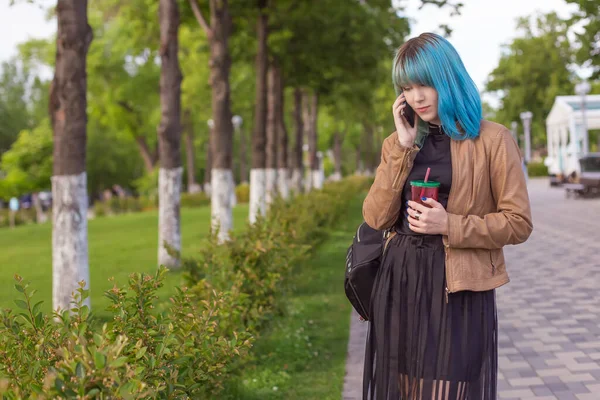 青い髪をした美しい若い女性は 街の公園の携帯電話で 彼女の手の中に飲み物とプラスチック製のガラスを話します ライフスタイル — ストック写真