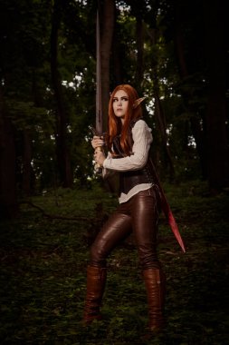 Fantezi kadın, keskin kulaklı elf savaşçısı, turuncu saçlı, deri korseli ve kılıçlı. Ormanda duruyor ve saldırmak için silah, savaş pozisyonu, savaş. KostümComment.