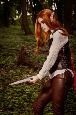 Fantezi kadın, keskin kulaklı elf savaşçısı, turuncu saçlı, deri korseli ve kılıçlı. Ormanda duruyor ve silahlarını keskinleştiriyor, Blade. Tehlikeli bir kız..