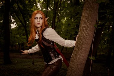 Güzel bir elf kadın, yaz akşamı ormanında bir savaşçı, bir ağaç sapının arkasından izliyor. Bir avcı, turuncu saçlı bir şef. KostümComment.