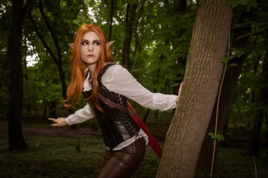 Güzel, genç bir kadın elf, keskin kulaklı bir savaşçı, ormanda, bir ağaç sapının arkasından izliyor. Bir avcı, turuncu saçlı ve deri korseli bir şef. KostümComment.