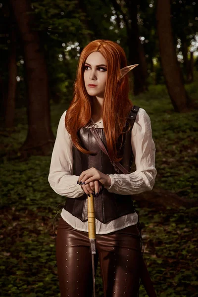 Fantasy woman, elf warrior with sharp ears, with orange hair and leather corset and with a sword. It is in the forest. Role-playing game. Cosplay.