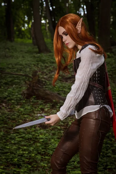 Fantasy woman, elf warrior with sharp ears, with orange hair and leather corset and with a sword. It stands in the woods and sharpens its weapons, blade. A dangerous fairy is a fighter.