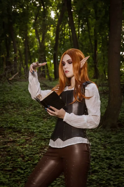 Fantasy woman, elf warrior with sharp ears, with orange hair and leather corset and with a sword. He stands in the woods, holds a magic book and utters a spell. A fiery ball. Cosplay.
