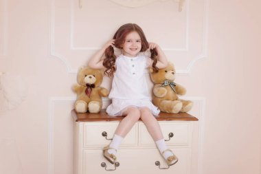 Odasında oyuncak dolu bir çekmecenin üzerinde oturan küçük bir kız. Beyaz elbiseli çocuk..