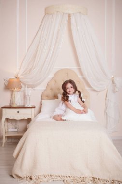Odasında küçük bir kız yatakta oturup bir yastığa sarılıyor. Beyaz elbiseli çocuk. İçi bej.