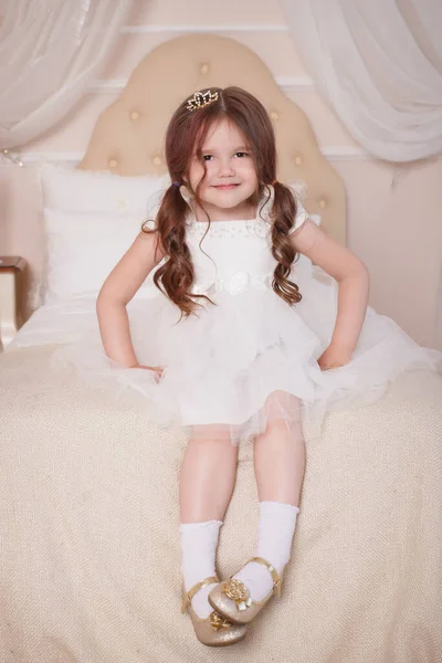 The child, the girl sits on a bed in a child's beige room, she is dressed in a white dress, and on her head is a caron. Little Princess.