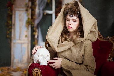 Bir prensesin ortaçağ kıyafeti, bir büyücü, kafasında başlık ve taç olan bir cadı. Antika kırmızı bir koltuğa oturuyor ve beyaz bir kafatası tutuyor. Güzel genç kız, kadınlar..