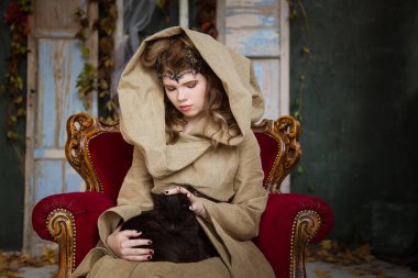 Bir prensesin ortaçağ kıyafeti, bir büyücü, kafasında başlık ve taç olan bir cadı. Antika kırmızı bir koltukta oturuyor ve kollarında siyah bir kedi tutuyor. Güzel genç kız, kadınlar..