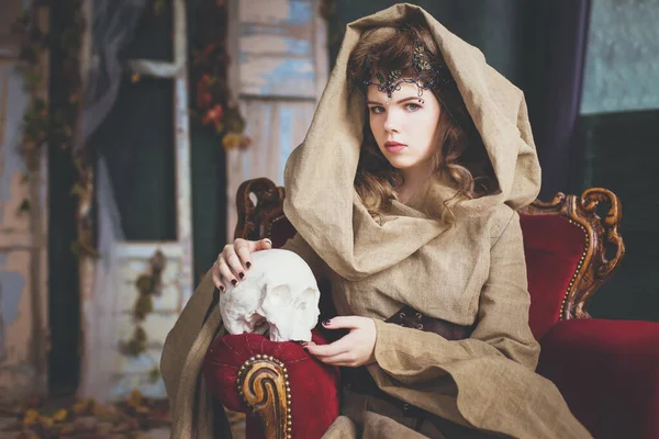 Medieval outfit of a princess, a sorceress, a witch with a hood and a tiara on her head. She sits on an antique red armchair and holds a plaster, white skull. Beautiful girl teenager, women.