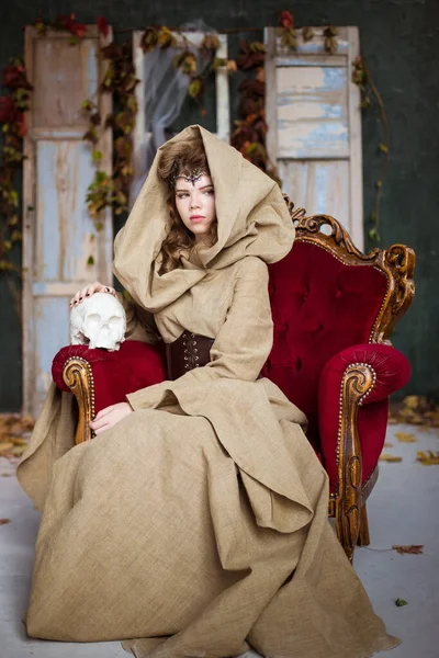 Medieval outfit of a princess, a sorceress, a witch with a hood and a tiara on her head. She sits on an antique red armchair and holds a plaster, white skull. Beautiful girl teenager, women.