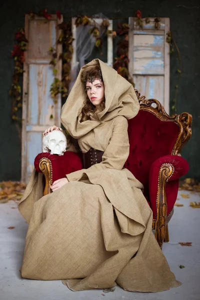 Medieval outfit of a princess, a sorceress, a witch with a hood and a tiara on her head. She sits on an antique red armchair and holds a plaster, white skull. Beautiful girl teenager, women.