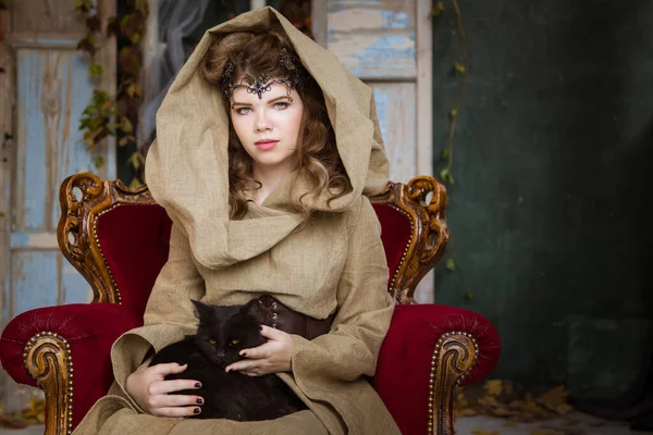 Medieval outfit of a princess, a sorceress, a witch with a hood and a tiara on her head. She sits on an antique red armchair and holds a black cat in her arms. Beautiful girl teenager, women.