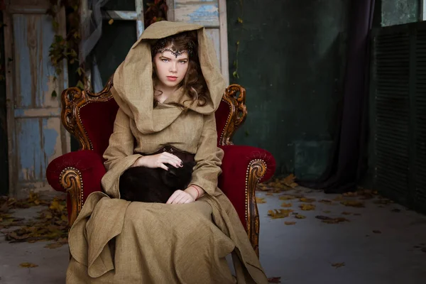 Medieval outfit of a princess, a sorceress, a witch with a hood and a tiara on her head. She sits on an antique red armchair and holds a black cat in her arms. Beautiful girl teenager, women.
