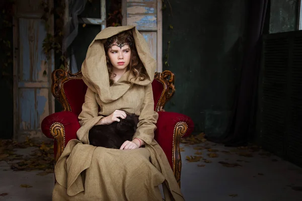 Medieval outfit of a princess, a sorceress, a witch with a hood and a tiara on her head. She sits on an antique red armchair and holds a black cat in her arms. Beautiful girl teenager, women.