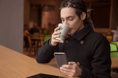 Rasta ve siyah ceketli yakışıklı genç bir adam akıllı telefona bakar ve kağıt bardaktan su içer. Kahve molası, kahvaltı, halka açık bir yer. Haberleri, sosyal ağları, interneti oku.