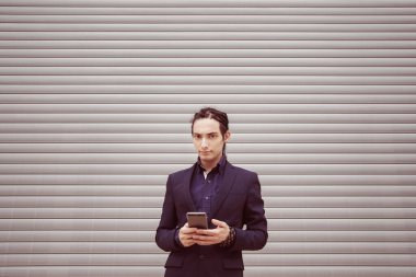 Rastalı ve takım elbiseli yakışıklı bir genç elinde akıllı telefonunu tutuyor, interneti ve sosyal ağları araştırıyor. Bir paten panjurunun arka planına karşı.