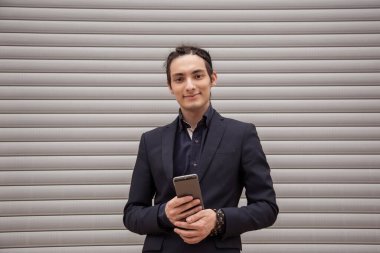 Rastalı ve takım elbiseli yakışıklı bir genç elinde akıllı telefonunu tutuyor, interneti ve sosyal ağları araştırıyor. Bir paten panjurunun arka planına karşı.