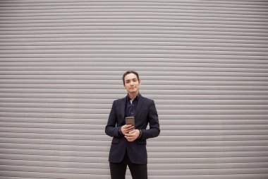 Rastalı ve takım elbiseli yakışıklı bir genç elinde akıllı telefonunu tutuyor, interneti ve sosyal ağları araştırıyor. Bir paten panjurunun arka planına karşı.