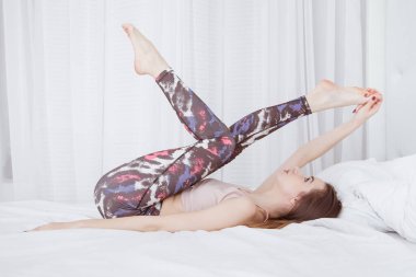 Genç bir kadın yatakta sırt üstü uzanıyor ve gerinme egzersizleri yapıyor. Yataktan kalkmadan yoga dersleri. İyi sabahlar. Beyaz arkaplan, iç plan.