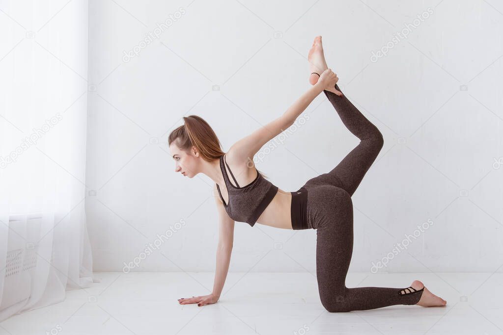 Fit young Caucasian woman practicing yoga at home standing in cat and cow pose arching her back. Raise your leg above your head. Lifestyle.