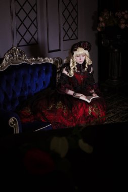 Güzel, küçük, sarışın bir kız kırmızı, gür, eski bir elbiseyle pahalı bir koltukta oturuyor. Kitap oku. Kadın, vampir Claudia. Bir prenses. Japon usulü lolita, güzel kostüm. Sokak modası.