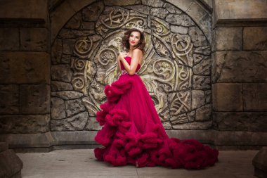 Güzel bir kadın, kırmızı lüks elbiseli bir kraliçe, orta çağ arkaplanında duruyor, kemerli bir Gotik amblem, kristalli gümüş karton. Kaledeki Prenses.