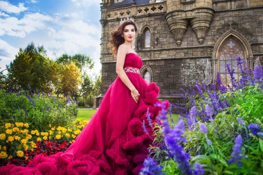 Güzel bir kadın, bordo elbiseli bir kraliçe, çiçek açan bir bahçede yürüyor. Arka planda antik bir kale.