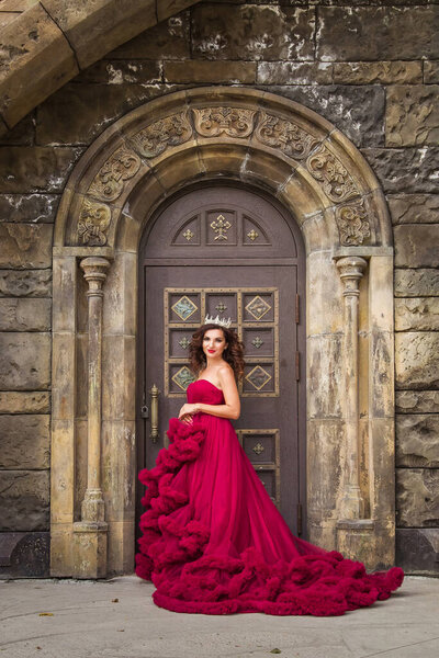 Красивая женщина, королева в красном роскошном платье, стоит на фоне средневековой, средневековой, готической двери с аркой. Средневековые фантазии. Принцесса

