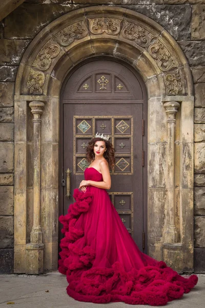 美しい女性 赤い豪華なドレスの女王は アーチを持つゴシック様式のドアを背景に立っています 中世のファンタジー 魅力的なプリンセス — ストック写真
