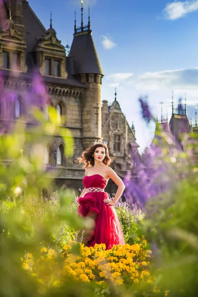 赤い豪華なドレスを着た美しい女性が咲く庭に立っています 背景にある古代の城 — ストック写真