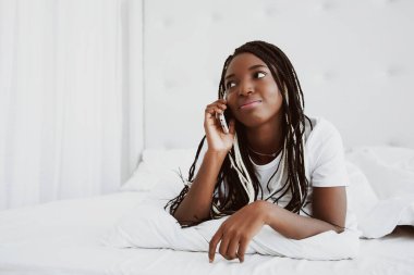 Güzel siyah bir kadın beyaz bir yatakta uzanıyor ve telefonda konuşuyor. Günaydın, bir arkadaşla iletişim..