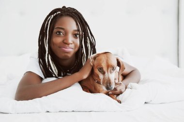Güzel bir Afrikalı kadın, kırmızı köpek dachshund ile bir yatakta, evcil bir evcil hayvanla birlikte yüzüstü yatıyor. İyi sabahlar. Portre. Nijerya, Afrika.
