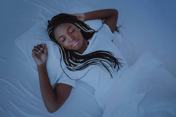 A beautiful African woman lies and sleeps in bed, in a white bedroom. Good night, vacation at home. The girl is wearing a T-shirt, pigtails on her head. Healthy sleep. Nigeria, Africa.