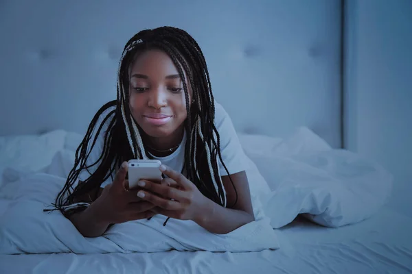 A beautiful African woman lies and bed, in a white bedroom and keeps the smartphone. Good night, vacation at home. The girl is wearing a T-shirt, pigtails on her head. Healthy sleep. Nigeria, Africa.