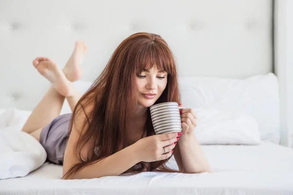朝のコーヒー ココア 紅茶のマグカップとベッドに座って美しい白人の女の子 白いベッドとインテリア グレーのパジャマナイトガウン アメリカ人 — ストック写真