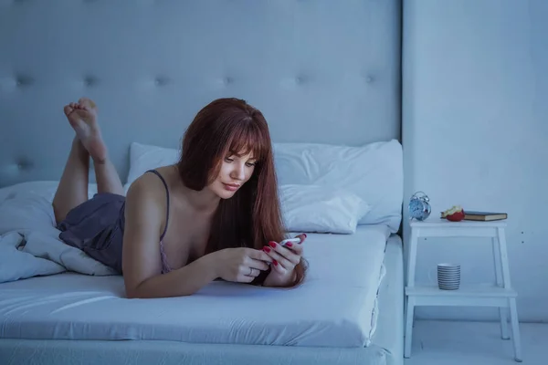 コミュニケーションと人々のライフスタイルのコンセプト 幸せな若い女性は 自宅の寝室でスマートフォンでベッドに横たわっています グレーの絹のパジャマ 夕暮れ — ストック写真