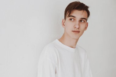 Beyaz bir duvara yaslanmış beyaz kolsuz tişörtlü genç ve yakışıklı bir adamın portresi. Öğrenci, yaşam tarzı. Boşluğu kopyala.