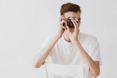 Yakışıklı bir genç adam, Sovyet, eski ayna kamerasıyla fotoğraf çekiyor ve dirseklerini sandalyenin arkasına yaslıyor. Beyaz arka planda beyaz bir forma giymiş bir adam var..