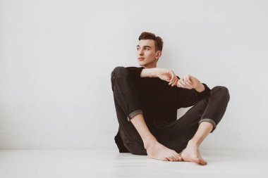 Genç bir adam, bir öğrenci yerde oturuyor, beyaz bir duvarın arkasında, dalgın dalgın bakıyor. Adam manken..