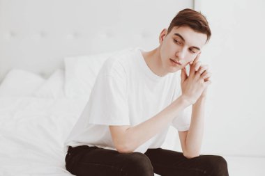 Beyaz tişörtlü, genç, yakışıklı bir adam yatağın köşesinde, yatak odasında oturuyor. Boşluğu kopyala.