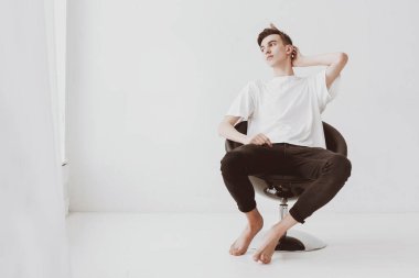 Düşünceli, sıradan elbiseli bir adam evinin penceresinin yanındaki bir sandalyede beyaz bir duvara karşı oturuyor. Boşluğu kopyala.