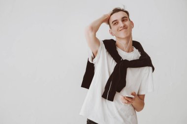 Beyaz arka planda oturan yakışıklı genç bir adam elinde telefon, kulaklık takıyor, müzik dinliyor ve saçını düzeltiyor, karizmatik bir adam. Boşluğu kopyala.