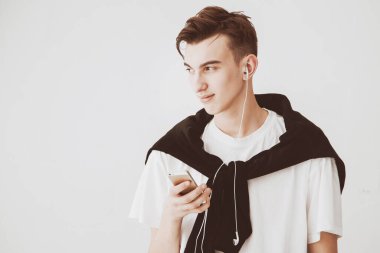 Beyaz arka plandaki yakışıklı genç adam elinde telefon, kulaklık takıyor ve müzik dinliyor. Boşluğu kopyala.