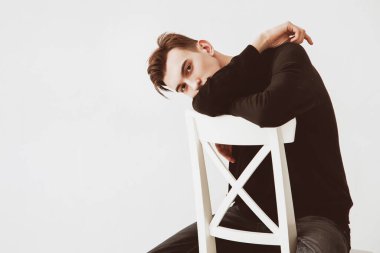 Siyah süveter giyen, beyaz sandalyede oturan güzel, genç bir adamın portresi..