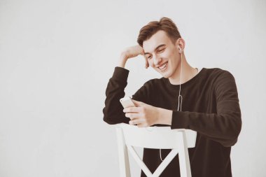 Genç bir adam, sandalyede oturan bir öğrenci, elinde bir telefon tutan ve kulaklıkla müzik dinleyen beyaz bir duvara karşı. Erkek model gülümsüyor..